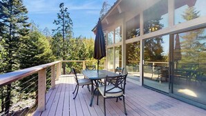 Un terrazzo in legno con tavolo e sedie, circondato da pareti di vetro e vista su alberi.