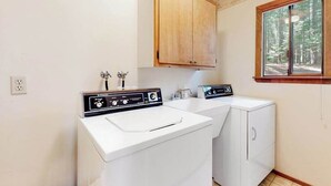 A laundry room with two washing machines, a wooden cabinet, and a window with a forest view. - Cresta Cabin - Pinebrook (Arnold)