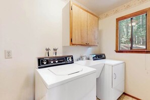 A laundry room with two washing machines, a wooden cabinet, and a window with a forest view. - Cresta Cabin - Pinebrook (Arnold)
