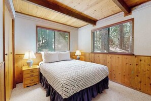 Ein Schlafzimmer mit Holzboden, zwei Nachttischlampen, einem Bett mit gemustertem Überwurf und Fenstern mit Blick auf einen Wald.