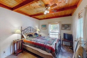 A bedroom with a wooden ceiling, a bed with a quilt, two bedside lamps, a television, and a window with curtains. - Casita Villareal - East Village (Shaver Lake)