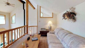 Et værelse med synlige træbjælker i loftet, en sofa, et bord med bøger og en lampe, samt et dekorativt vægkunstværk.