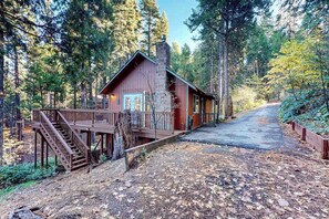 Eine Holzhütte mit einer Holzterrasse und einer Treppe, die zu einer Tür führt, umgeben von einem bewaldeten Gebiet.