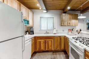 A kitchen with wooden cabinets, a white refrigerator, a stove, and a window with blinds. - Snowpeak Chalet - Big Trees Village (Camp Connell)