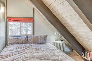 A bedroom with a sloped ceiling, a bed with a plaid bedspread, a window with a red valance, and a small table with a lamp and books.