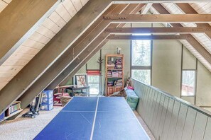 An attic room with a ping pong table, wooden beams, a bookshelf, and a window.