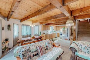 Een ruime woonkamer met een houten plafond, een bank met gestreepte kussens, een eettafel en een keuken op de achtergrond.