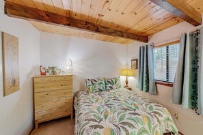 Een slaapkamer met een houten plafond, een bed met een beddengoed met een patroon, een dressoir, een bedlamp en een raam met gordijnen.