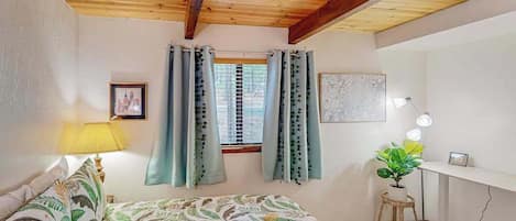 Un dormitorio con cama, escritorio, lámpara, ventana con cortinas e imagen colgada en la pared.