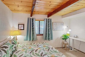 Un dormitorio con cama, escritorio, lámpara, ventana con cortinas e imagen colgada en la pared.