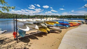 Una hilera de canoas y kayaks alineados en una playa arenosa junto a un lago tranquilo con cielo despejado.
