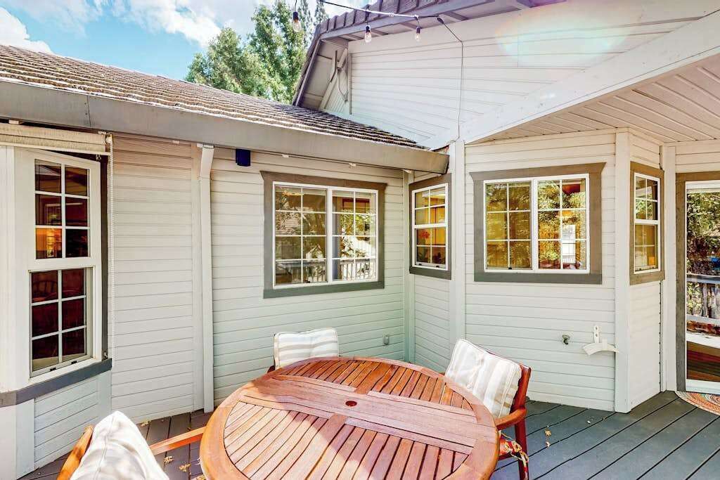 A covered patio with a wooden table and chairs, large windows, and a white exterior wall.