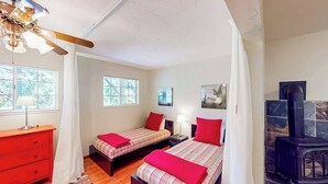 Un dormitorio con dos camas, un aparador rojo, un ventilador de techo de madera, una chimenea de piedra y una ventana con vista a árboles.