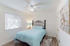 A bedroom with a bed, a ceiling fan, a window with blinds, a nightstand with a lamp, and a wall with a painting. - Holiday Chalet - Pine Mountain Lake (Groveland)