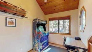 A room with a wooden ceiling, a window with a view of trees, a table with a chair, and a colorful arcade machine. - Hot Tub Hideaway - Big Trees Village (Dorrington)