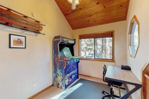 A room with a wooden ceiling, a window with a view of trees, a table with a chair, and a colorful arcade machine. - Hot Tub Hideaway - Big Trees Village (Dorrington)
