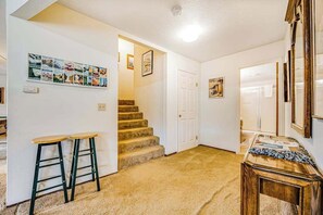 Ein Flur mit Teppichboden, einer Treppe und einer Tür, die in einen anderen Raum führt. Zwei Hocker und ein kleiner Tisch mit Papieren darauf sind ebenfalls zu sehen.