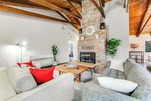Ein Wohnzimmer mit Kamin aus Backstein, Holzbalken und einem weißen Sofa mit roten und weißen Kissen.