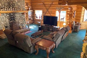 A log cabin interior with a stone fireplace, leather sofas, a flat-screen TV, and a wooden entertainment center.
