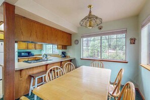 A kitchen with wooden cabinets, a dining table, and chairs. There is a window with a view of trees. - Traveler's Rest - East Village (Shaver Lake)