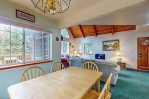 A dining area with a wooden table and chairs, a sofa, and a television in a room with wooden beams and large windows. - Traveler's Rest - East Village (Shaver Lake)