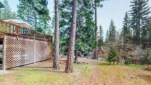 A wooden deck with a lattice fence, surrounded by tall trees and a grassy area. - The Dollhouse Cabin - Blue Lake Springs (Arnold)