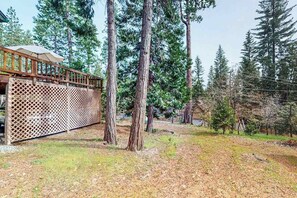 A wooden deck with a lattice fence, surrounded by tall trees and a grassy area. - The Dollhouse Cabin - Blue Lake Springs (Arnold)