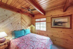 A wooden cabin bedroom with a bed, a window, a ceiling fan, and a painting on the wall. - The Dollhouse Cabin - Blue Lake Springs (Arnold)