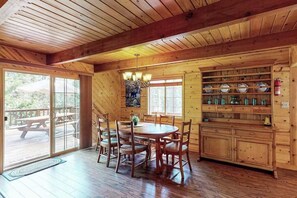 Un interior de madera con una mesa de comedor, sillas, una araña y una zona de cocina con estantes.