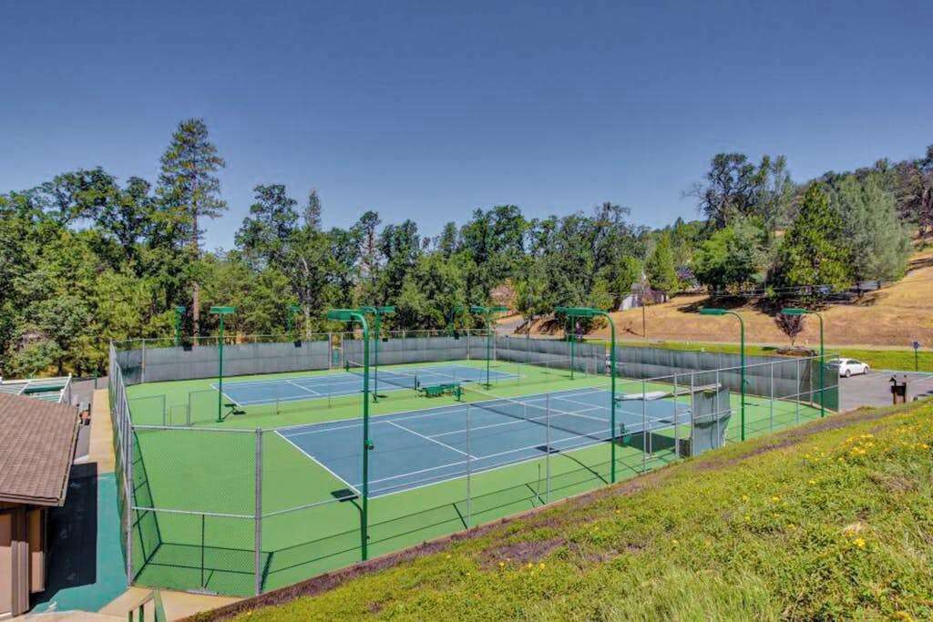 A tennis court with a surrounding fence, trees, and a hilly landscape.
