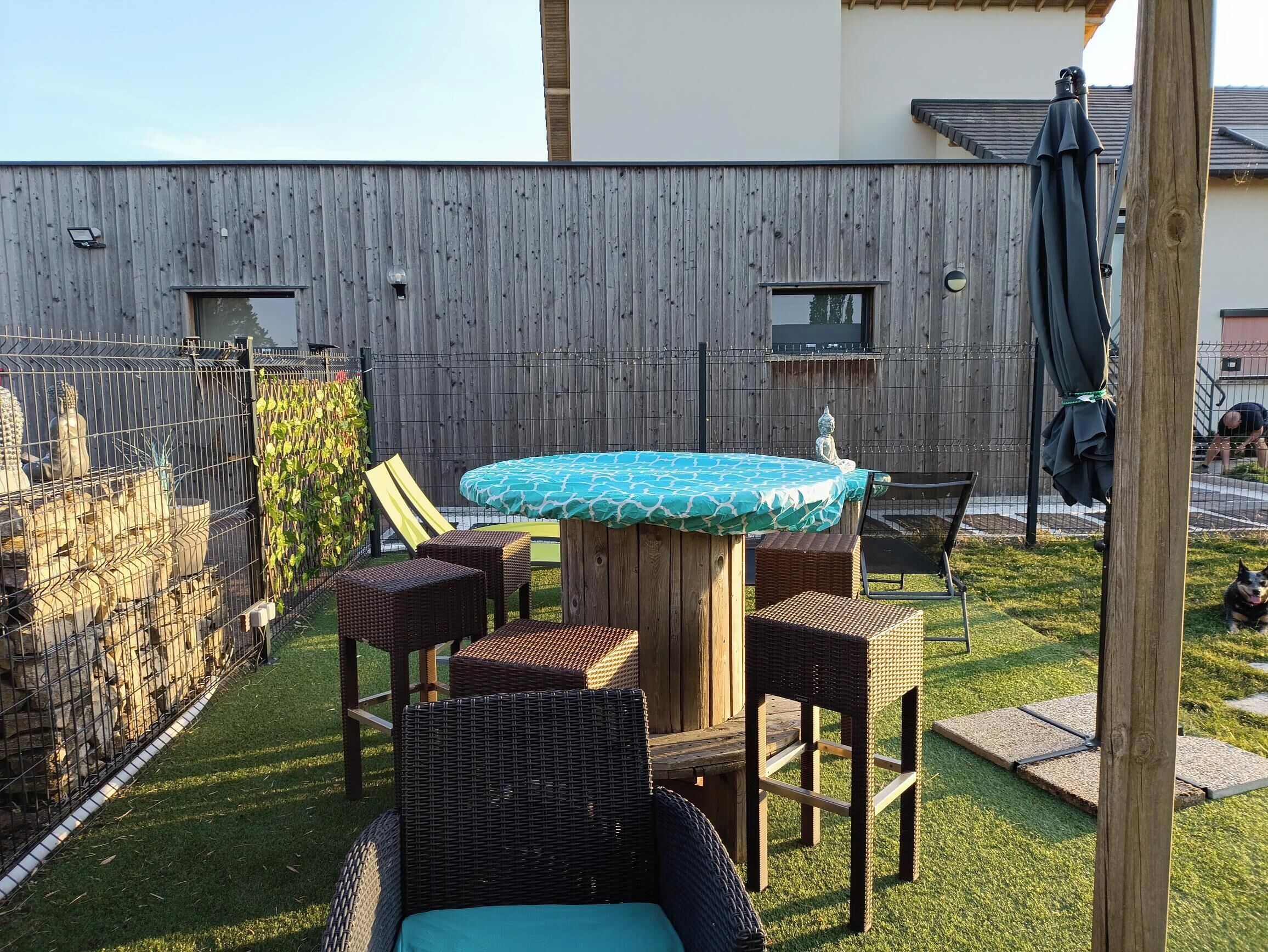 Un jardin avec une table ronde, quatre chaises en osier, une petite statue et une clôture en bois.