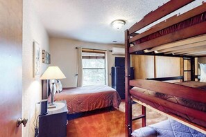 Une chambre avec des lits superposés, une table de chevet avec une lampe, une fenêtre avec des stores et un luminaire au plafond.