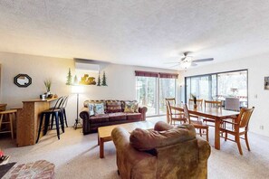 A spacious living room with a large window, a ceiling fan, and a dining area with wooden furniture. - Creekside Cabin - Pine Mountain Lake (Groveland)