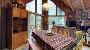 Een gezellig chaletinterieur met een eettafel, houten stoelen, een bankje, een open haard en een groot raam.