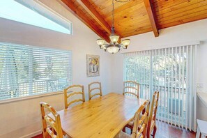 Un coin repas avec une table et des chaises en bois, un lustre et de grandes fenêtres équipées de stores.