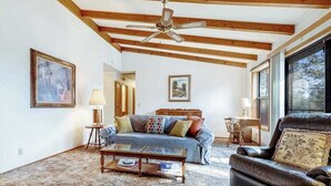 Ein Wohnzimmer mit einer Couch, einem Couchtisch und einem Ledersessel. Es gibt einen Deckenventilator, Holzbalken und ein großes Fenster.