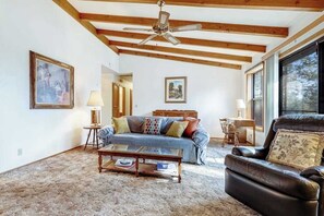 Ein Wohnzimmer mit einer Couch, einem Couchtisch und einem Ledersessel. Es gibt einen Deckenventilator, Holzbalken und ein großes Fenster.