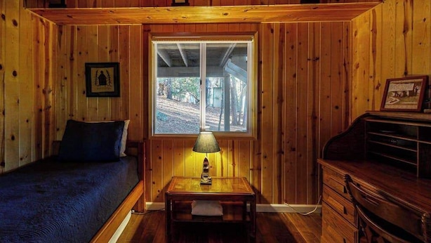 Una habitación tipo cabaña de madera con una cama, una mesa con una lámpara, un ventanal y un aparador.