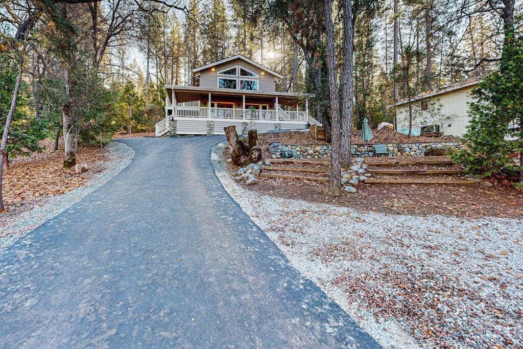 A two-story cabin with a porch, surrounded by trees and a gravel driveway.