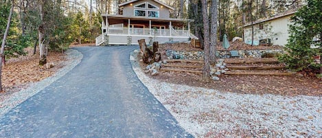 A two-story cabin with a porch, surrounded by trees and a gravel driveway.