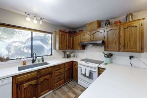 A kitchen with wooden cabinets, a white countertop, a stove, and a window with a view of trees. - Top Deck Retreat - Huntington Lake (Lakeshore)