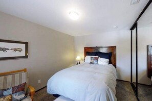A hotel room with a bed, a nightstand, a lamp, a framed picture on the wall, and a window. - Top Deck Retreat - Huntington Lake (Lakeshore)