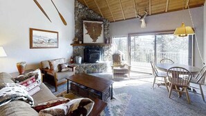 A cozy living room with a fireplace, a dining area with chairs, and a large window providing a view of the outdoors. - Top Deck Retreat - Huntington Lake (Lakeshore)