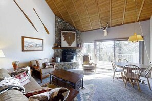 A cozy living room with a fireplace, a dining area with chairs, and a large window providing a view of the outdoors. - Top Deck Retreat - Huntington Lake (Lakeshore)