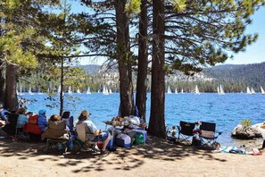 A lakeside picnic area with people seated on chairs, surrounded by trees and a view of sailboats on the water. - Weekend at the Lake - Huntington Lake (Lakeshore)