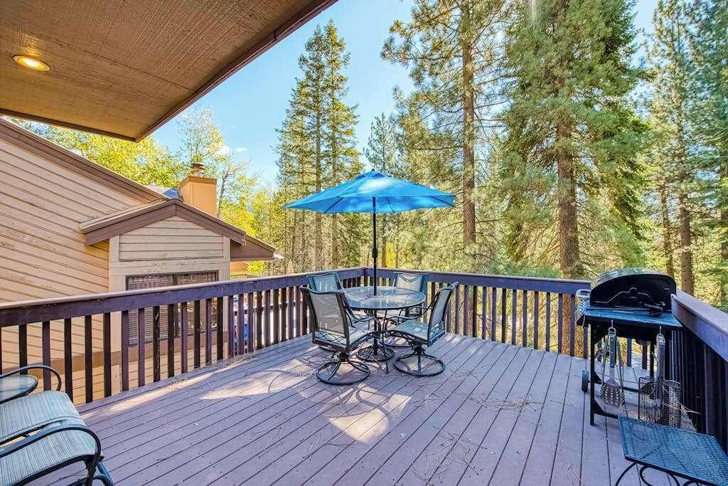 Um deck de madeira com mesa, cadeiras, churrasqueira e um guarda-sol azul.