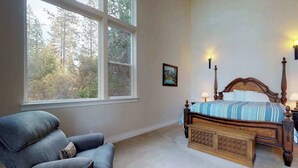 Ein Schlafzimmer mit großem Fenster, einem Bett mit Holz Kopfteil, einem blauen Sessel und zwei Wandleuchten.