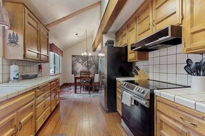 A kitchen with wooden cabinets, a stainless steel refrigerator, a stove, and a dining area with a table and chairs. - Whispering Pines - Sierra Cedars (Shaver Lake)