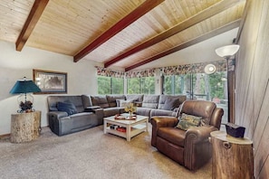 A living room with a sectional sofa, a leather armchair, a coffee table, and a lamp. There is a window with a view of trees and a framed picture on the wall. - Whispering Pines - Sierra Cedars (Shaver Lake)