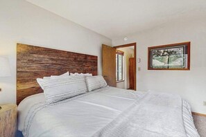 Un dormitorio con cabecera de madera, una cama grande con ropa blanca, un cuadro enmarcado en la pared y una puerta que da a otra habitación.